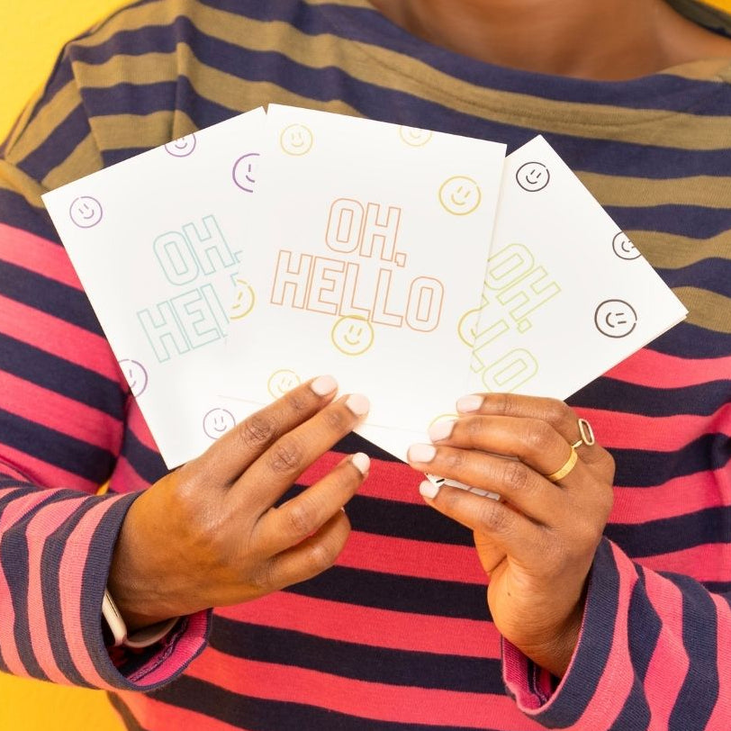 Person wearing a striped shirt holding 'OH HELLO' note cards against a yellow background. greeting cards